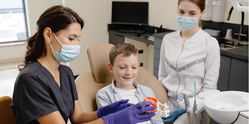 A kid sitting in a dental chair smiling a two dentist stand close by.